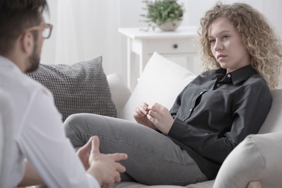 A young woman seated on a couch looks thoughtful as she listens to a therapist.