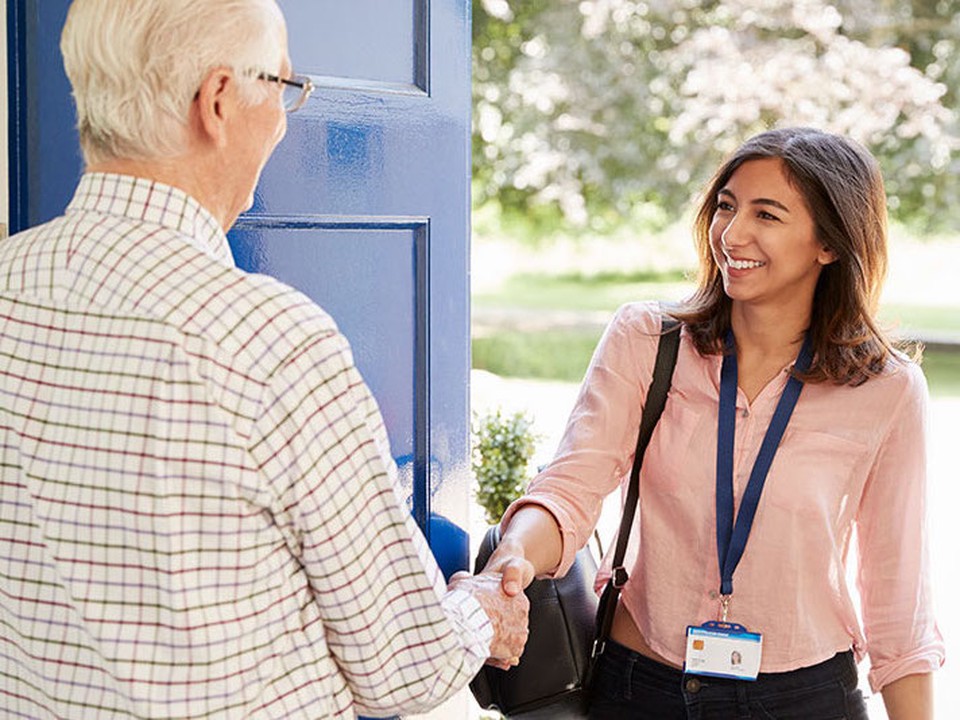 A female social worker greets an elderly man as she walks through his door. 