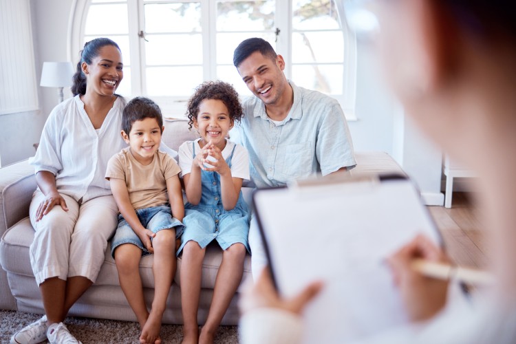 A social worker takes notes on a clipboard while interviewing a smiling family of four.