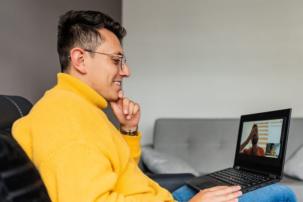 A man relaxing in a home environment smiles while participating in a video call on his laptop.