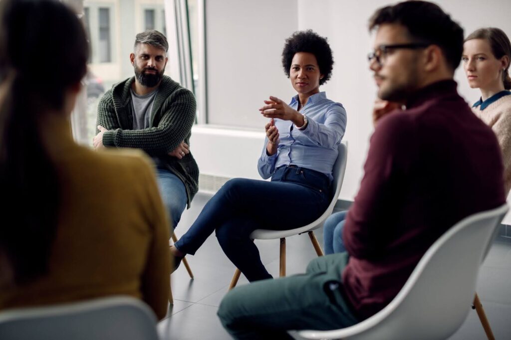 A woman speaks and gestures while leading a group session with four other adults seated in a circle in a bright, modern room.