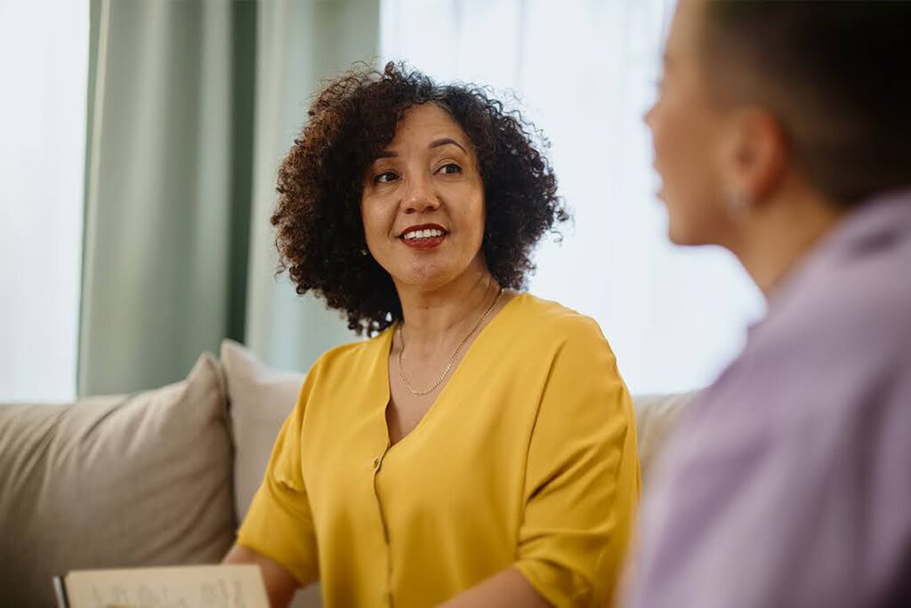A woman in a yellow blouse smiles and speaks with another person during a conversation; both sit on a couch in a bright, comfortable room.
