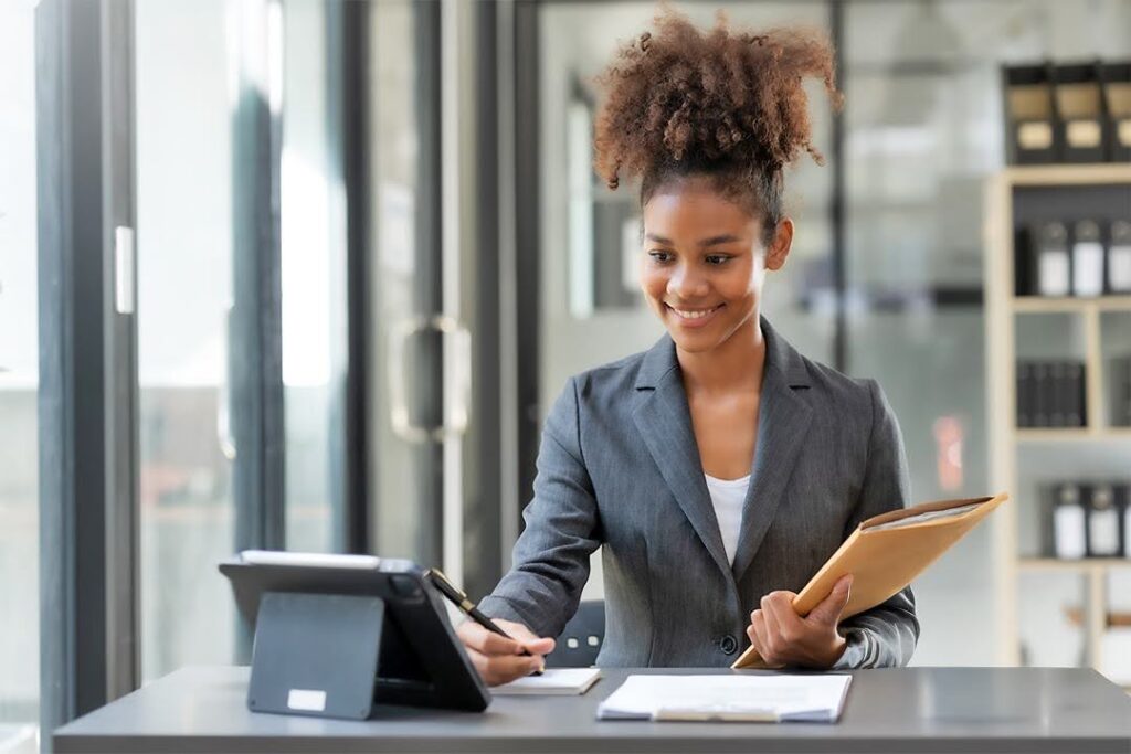 A woman in a blazer holds a folder and a pen and smiles at a tablet propped against her desk.